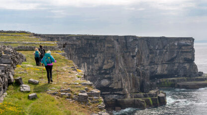 Three hikers with backpacks walk along a grassy cliff edge with steep rocky faces overlooking the ocean on a cloudy day, exploring Ireland's West Coast.