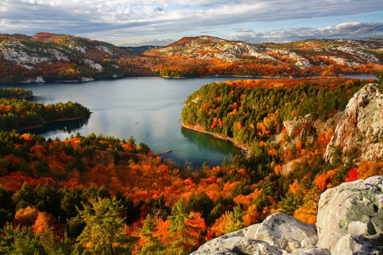 Experience a scenic view of a lake surrounded by rocky hills and dense forests, showcasing vibrant autumn foliage under a partly cloudy sky. This breathtaking scene is reminiscent of the beauty one might find when traveling through Ireland's enchanting landscapes.