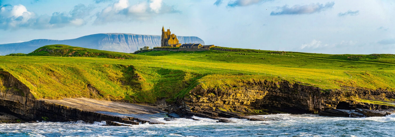 A castle majestically sits atop a lush green cliff in Ireland, overlooking a rocky shoreline with crashing waves. The distant landscape includes hills under a partly cloudy sky, making it one of the best regions to visit.