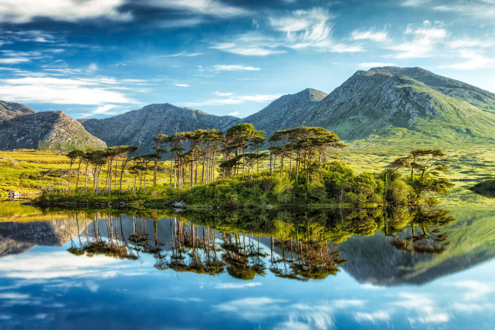 A small island with trees is reflected in a calm lake, set against a backdrop of green mountains under a blue sky with wispy clouds. It's an idyllic scene reminiscent of the serene landscapes one might encounter when traveling through Ireland.