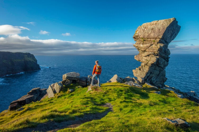 A person with a backpack stands on a grassy cliff overlooking the ocean in Ireland, near a large, uniquely shaped rock formation under a blue sky. Perfect for Ireland travel enthusiasts, this scene captures the essence of exploring scenic vistas at their best.