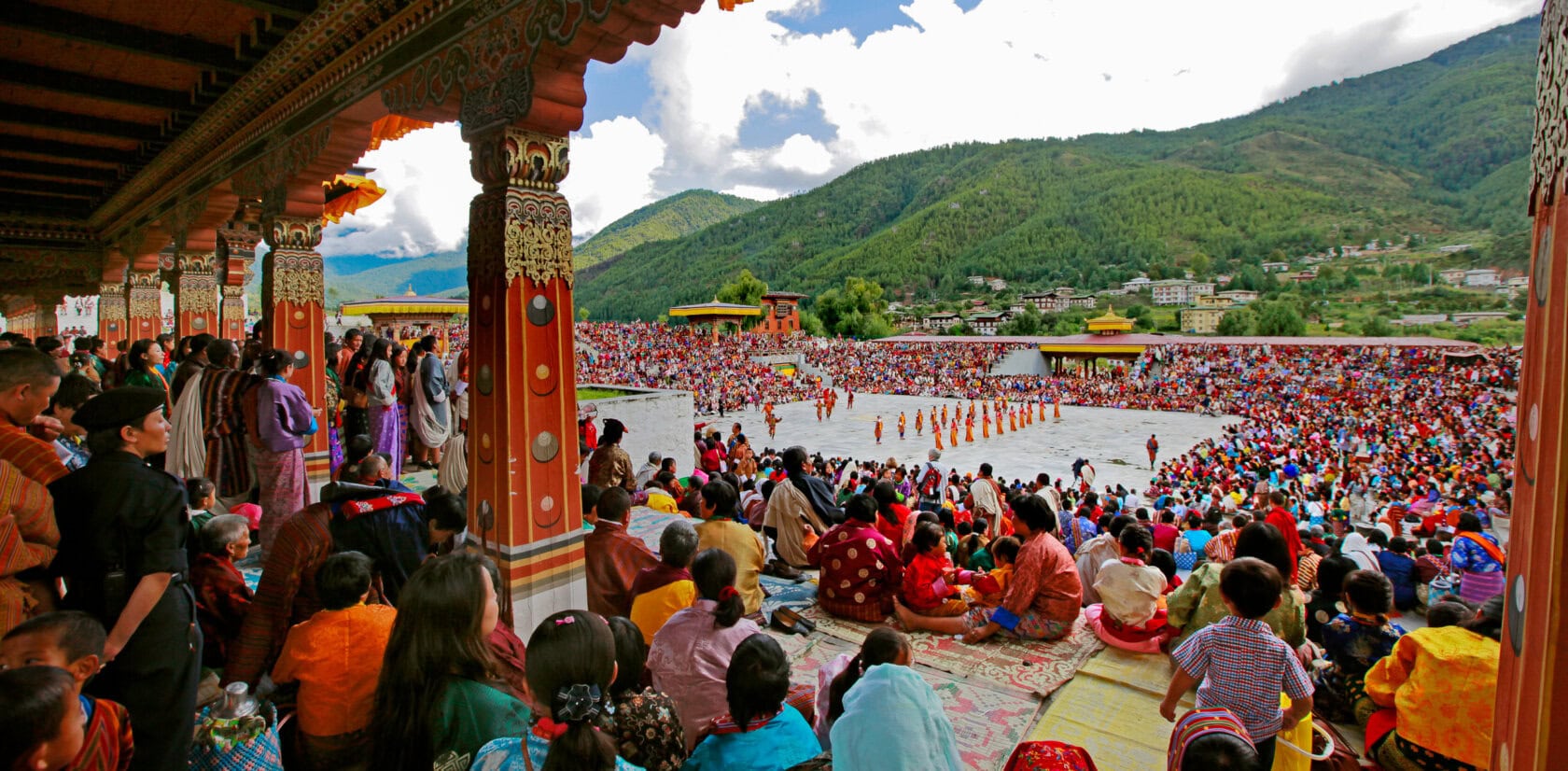 A large crowd watches a vibrant cultural performance, reminiscent of Bhutan's lively Tshechu festivals, in an open area surrounded by mountains. People are seated on a decorated balcony and the ground, fully immersed in the joyous event.