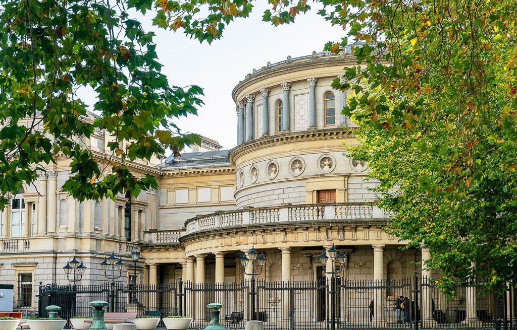 In Dublin, a historic stone building with large round towers and arched windows peeks through the trees, surrounded by a metal fence—a must-see for any travel itinerary.