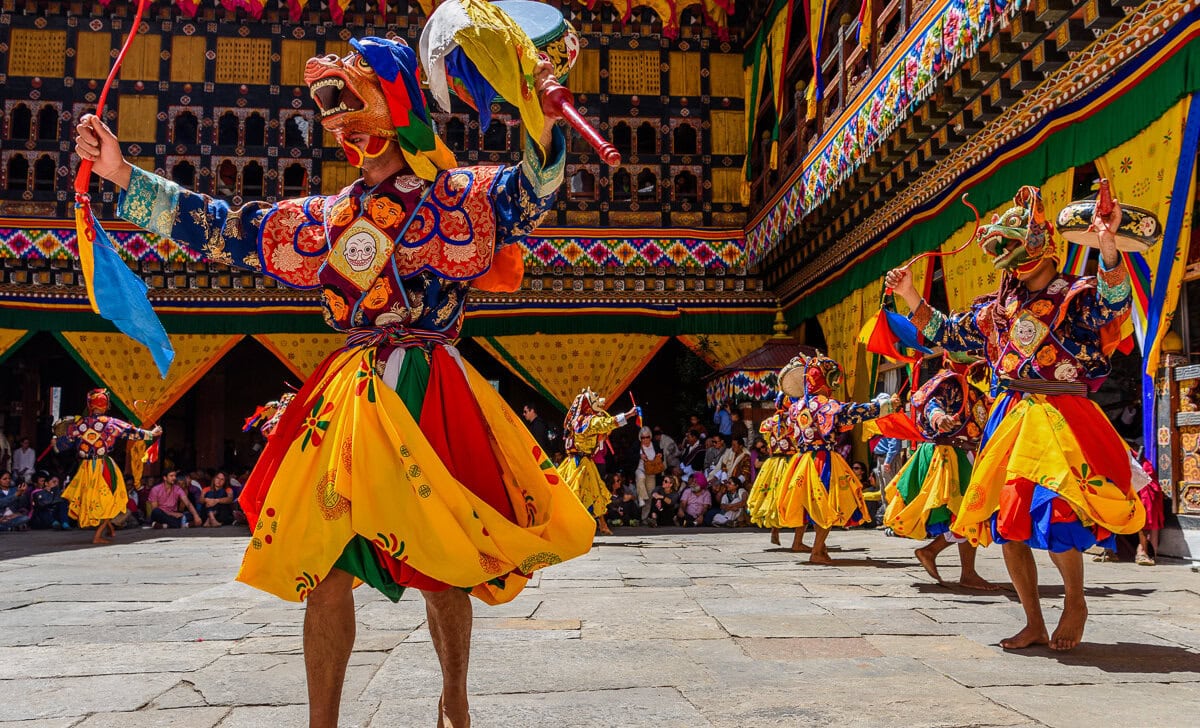 In a vibrant courtyard, colorfully dressed dancers donning masks perform a traditional dance as part of Bhutan's renowned Tshechu festival, captivating an eager audience with their rhythmic movements and vivid costumes.