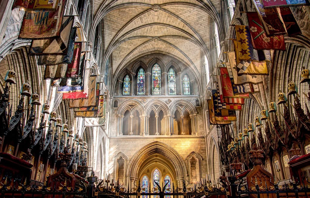 Explore the interior of a gothic cathedral in Dublin, showcasing high vaulted ceilings, stunning stained glass windows, and colorful flags adorning the upper walls—truly a must-see destination for travel enthusiasts seeking captivating things to do.