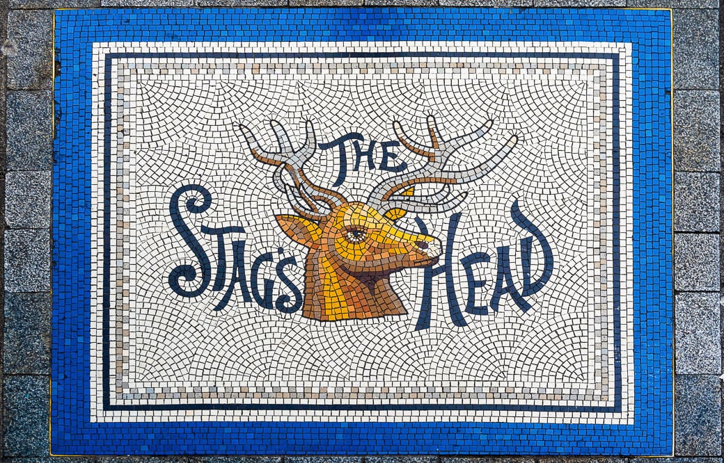 A mosaic artwork depicting a stag's head with "The Stags Head" text is framed by a blue border on a tiled surface, adding charm to one of Dublin's unique spots. Perfect for those exploring the city's vibrant art scene and seeking memorable things to do.