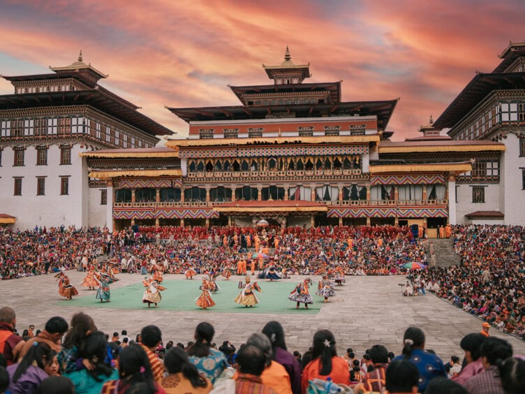A vibrant Tshechu festival unfolds in Bhutan, with dancers in colorful attire performing before a large crowd at a traditional architectural site during sunset.