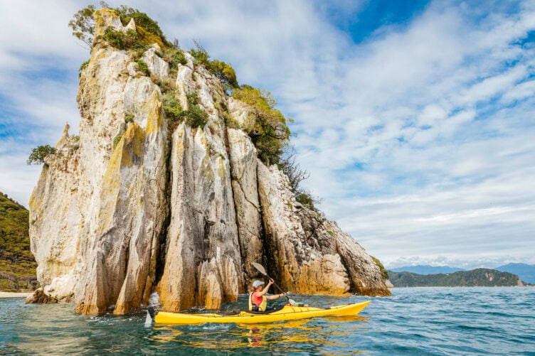 Experience the thrill of kayaking on the ocean near a large rocky cliff under a partly cloudy sky—one of the must-do adventures when exploring New Zealand's stunning landscapes.