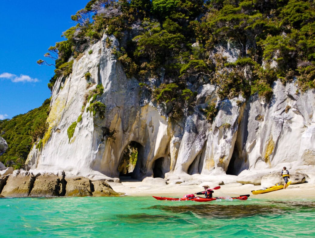 Two people kayaking near a rocky, forested coastline with caves in New Zealand's stunning national parks. The water is clear and turquoise, and the sky is a brilliant blue.