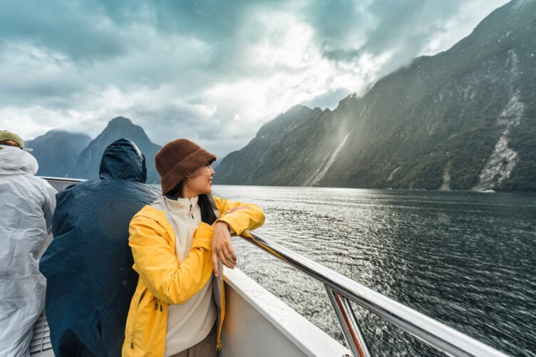 Adventurer in a yellow jacket and brown hat stands on a boat, gazing at New Zealand's breathtaking mountain landscape over the water. Nearby, fellow travelers in raincoats enjoy the journey under the cloudy sky—a perfect travel experience.