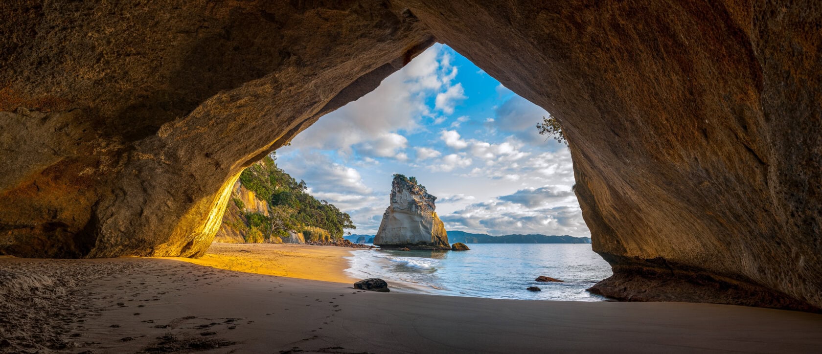 View from inside a large cave opening to a beach and clear blue sky in New Zealand. Sunlight illuminates the sandy floor and rock walls. Discover why this is the best island, with its isolated rock formation standing majestically in the sea, inviting you to visit.