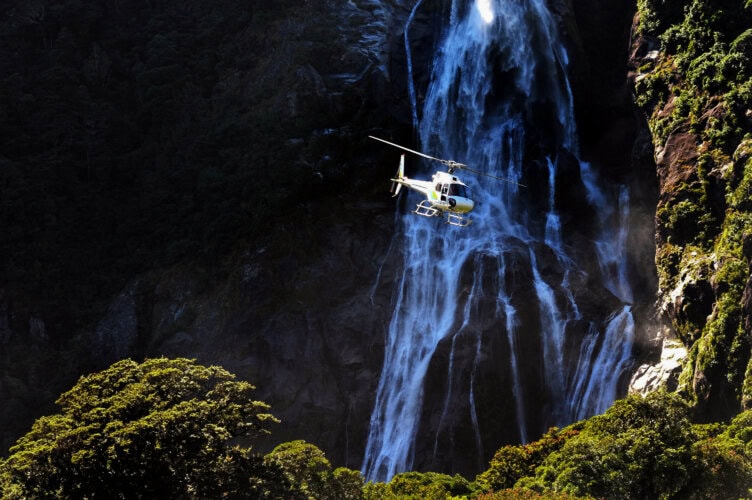 A helicopter soars in front of a majestic waterfall in New Zealand, surrounded by lush green foliage.