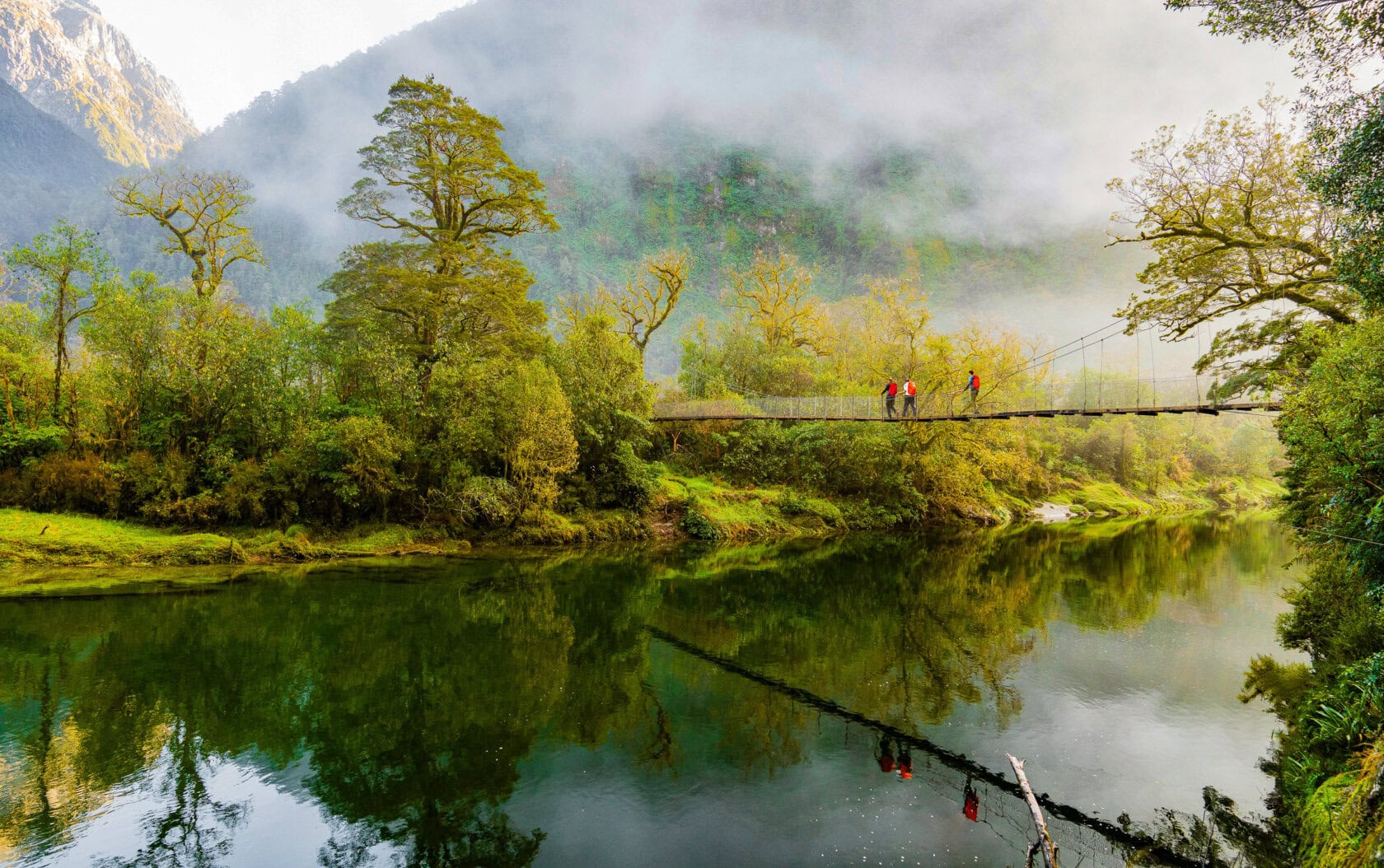 A lush river landscape with a suspension footbridge, where four people are walking. Misty mountains and dense greenery are reflected in the calm water—a perfect snapshot of a New Zealand trip.
