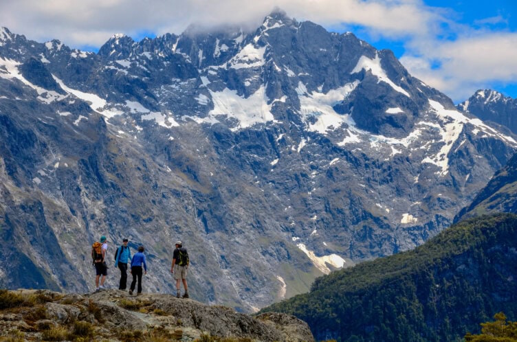 Four hikers stand on a rocky ledge, gazing at the tall, snow-capped mountains of New Zealand under a partly cloudy sky.