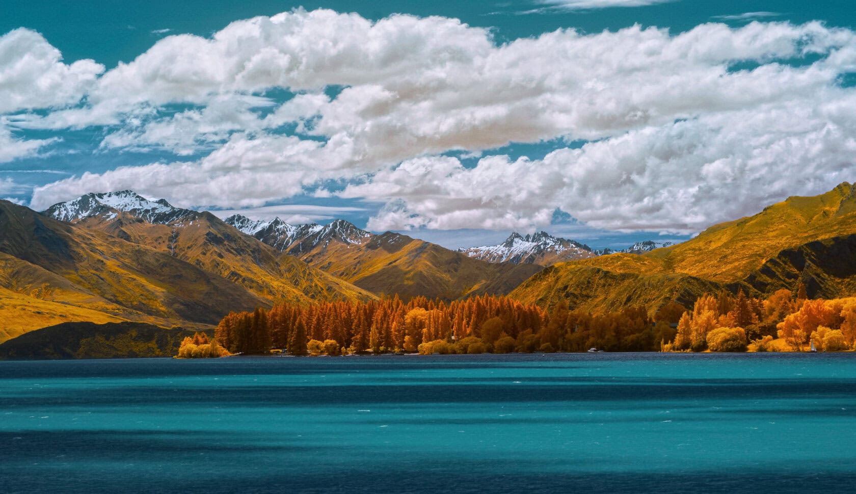 Discover a stunning lake with vibrant blue water, nestled amid autumnal trees and snow-capped mountains under a partly cloudy sky—an enchanting travel destination in New Zealand. The best time to visit is during the fall when nature's colors are at their peak.