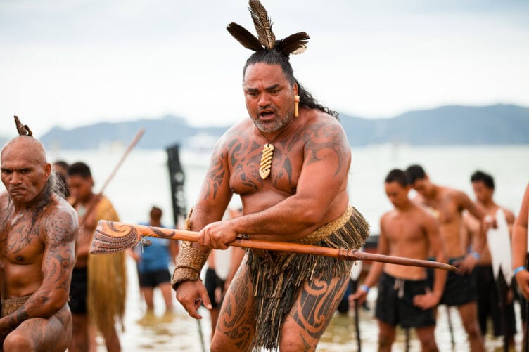 A group of people perform a traditional cultural dance on a beach in New Zealand, wearing traditional attire and holding wooden staffs. This captivating performance is one of the top activities to experience among things to do when visiting this beautiful country.