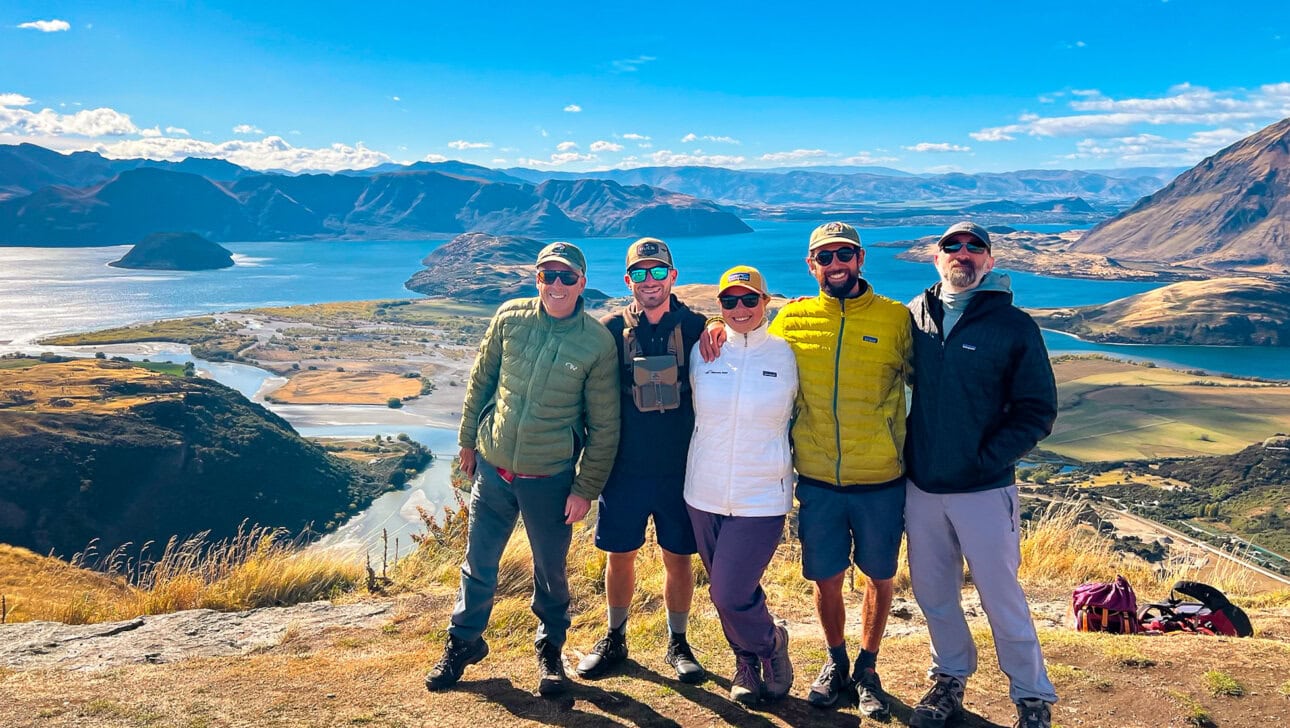 Five people wearing outdoor clothing stand on a hilltop, smiling at the camera, with a scenic view of lakes and mountains in the background.