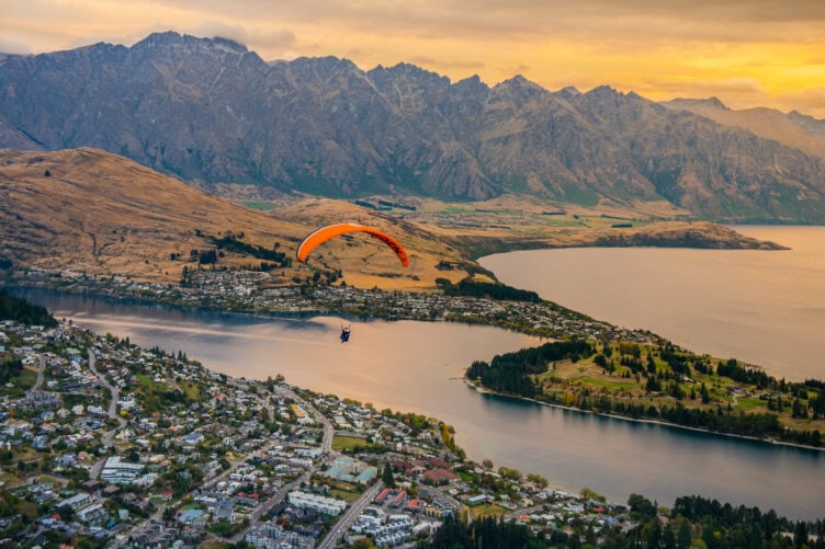 A paraglider with an orange canopy soars over a lake and town in New Zealand, with majestic mountains in the background under a cloudy sky at sunset. It's one of the unforgettable things to do for those who love travel and adventure.