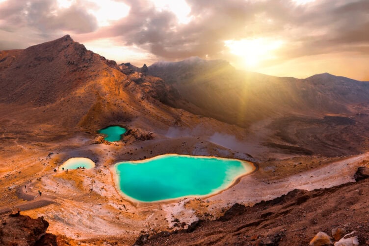 Discover the scenic beauty of New Zealand, where turquoise lakes shimmer amidst rugged, rocky terrain. Under a cloudy sky, sunlight gently peeks over distant mountains—a perfect addition to your list of things to do.