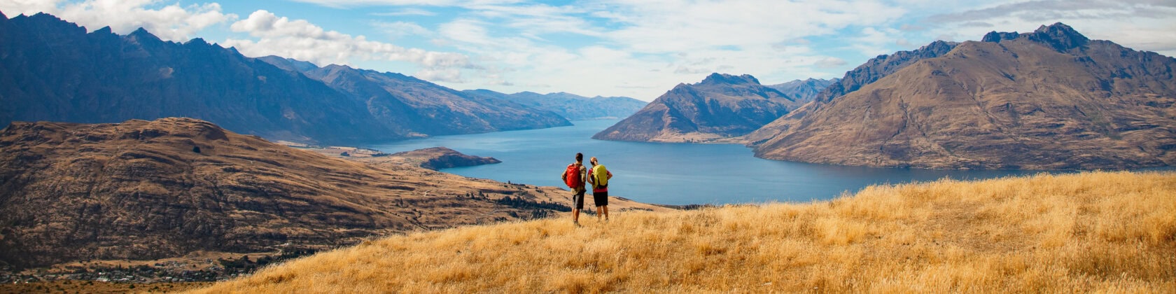 Two hikers stand on a grassy hill in New Zealand, overlooking a lake surrounded by majestic mountains under a blue sky. It's one of the top 15 things to do for nature lovers seeking breathtaking views and serene landscapes.