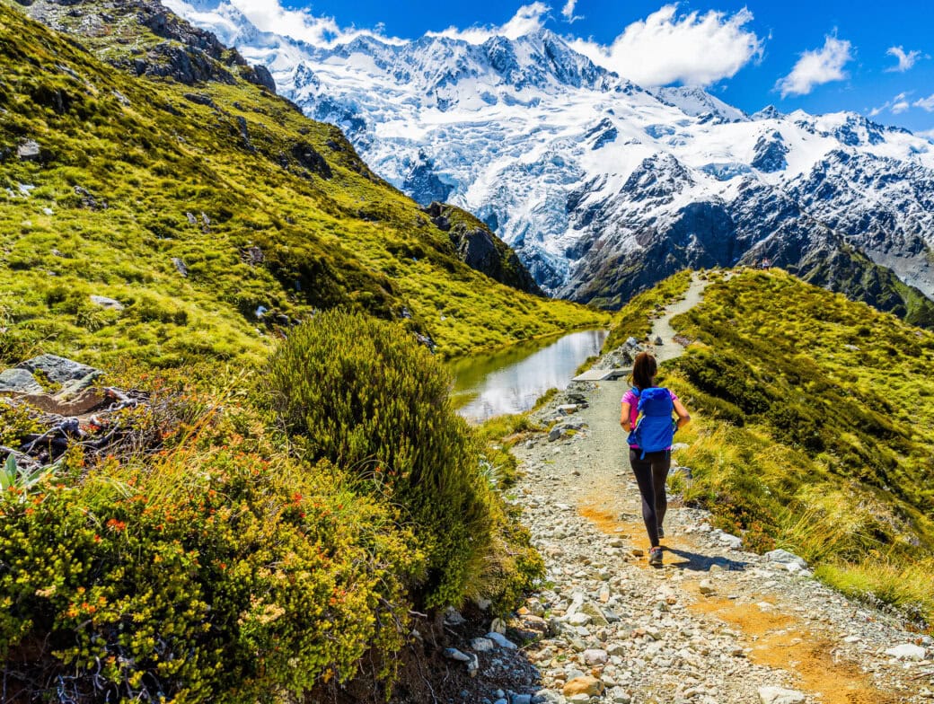A person hikes on a rocky trail through New Zealand's green hills, with snowy mountains painting the backdrop under a clear blue sky, showcasing the unparalleled beauty of its national parks.