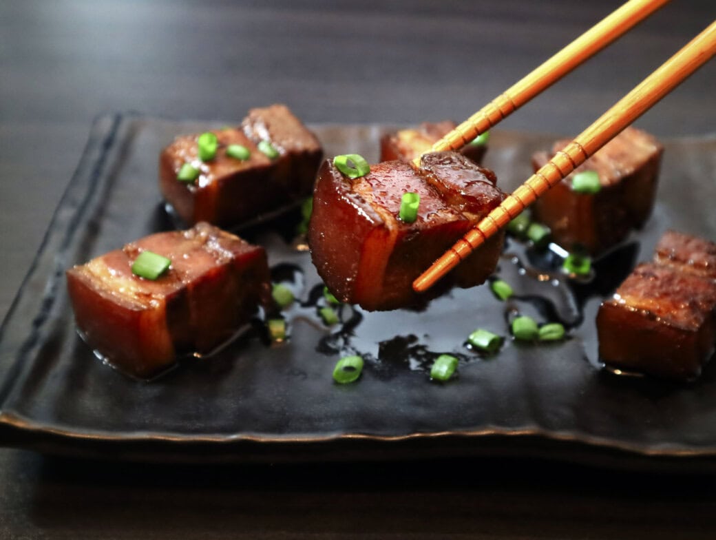 Chopsticks hold a piece of glazed meat garnished with chopped green onions on a black plate, with more pieces in the background—a culinary delight to savor after adventurous travel activities in Queenstown.