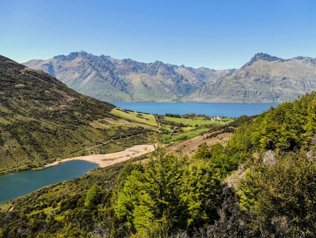Experience the scenic beauty of lush green hills and a serene lake, framed by jagged mountains under a clear blue sky—a perfect backdrop for your Queenstown travel adventures.
