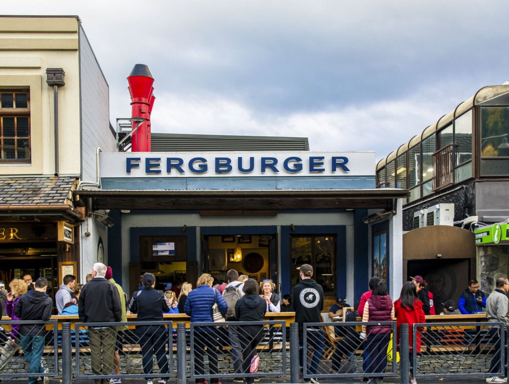 A crowd of people waits outside Fergburger, a popular Queenstown burger joint with a prominent red chimney and adjacent shops—a must-visit for any travel itinerary and one of the unforgettable things to do in town.