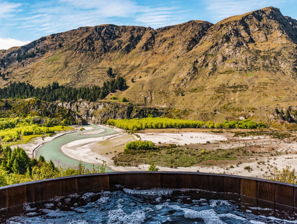 Soak in a hot tub while overlooking Queenstown's attractions—a winding river, vibrant green and yellow trees, and rocky mountains under a clear blue sky.