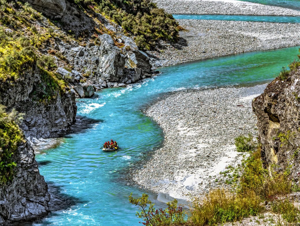 A group of people whitewater rafting down a winding turquoise river in Queenstown, surrounded by rocky cliffs and greenery, is one of the top attractions and thrilling things to do.