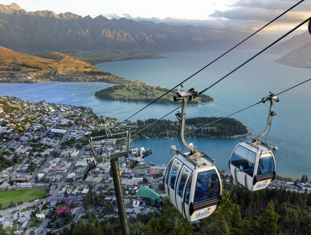 Two gondola cabins ascend a cable over Queenstown's stunning mountainous landscape. Beneath lies a charming town, a shimmering lake, and rolling hills, with majestic peaks in the background—a perfect vantage point for discovering top activities and exciting things to do.
