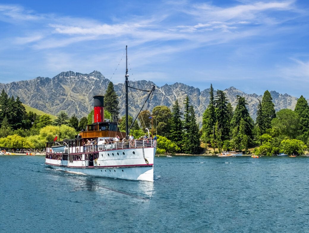 A steamship glides across the serene lake in Queenstown, framed by a mountainous backdrop and pine-lined shores under a clear blue sky, offering one of the most picturesque things to do on your travel itinerary.