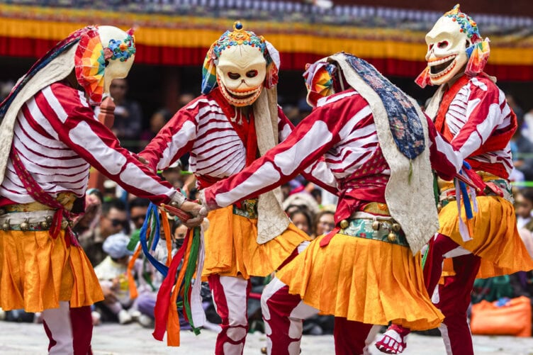 Four dancers in colorful skeleton costumes and masks perform in a circle, captivating the audience in the backdrop—a mesmerizing scene that travelers might stumble upon during their India travel adventures.