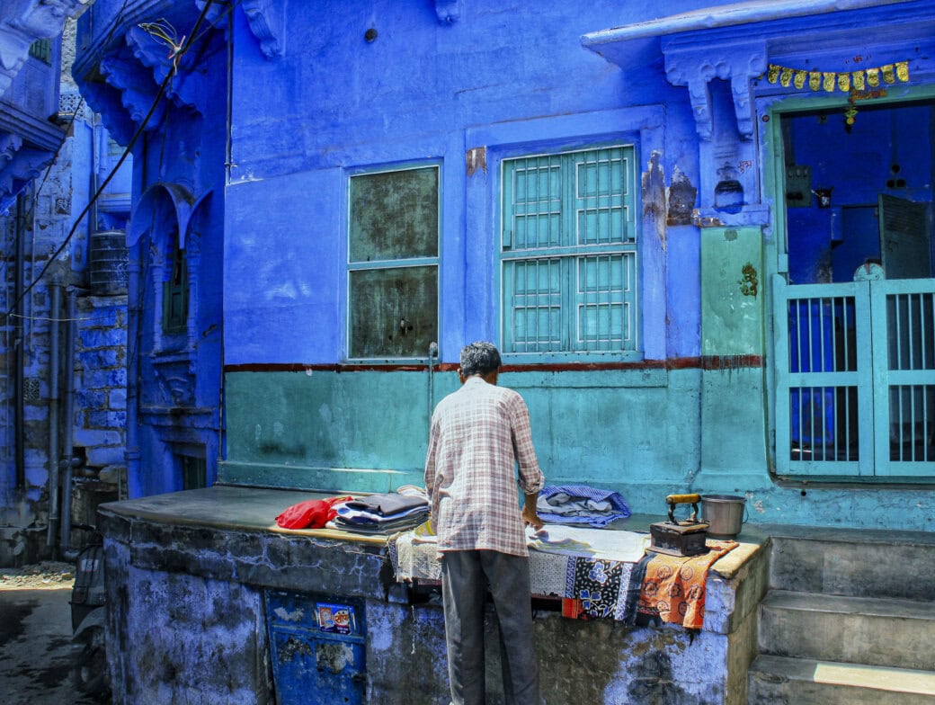 A person stands in front of a vibrant blue building in Rajasthan, India, arranging clothes on a table outside. The building features a small window and a door with bars, offering a colorful glimpse into the everyday life of this culturally rich region.