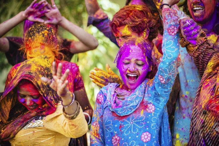 A group of people joyfully covered in colorful powder celebrate outdoors, with some raising their hands, capturing the vibrant spirit of Indian seasons.