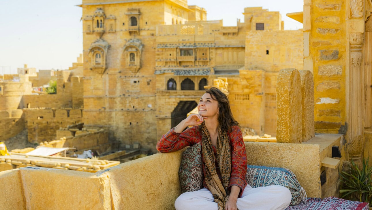 A person sitting cross-legged on a cushioned terrace in Rajasthan, looking up thoughtfully. Historic sandstone buildings rise majestically in the background under a clear sky, making this moment feel like it belongs in a beginner's guide to India.