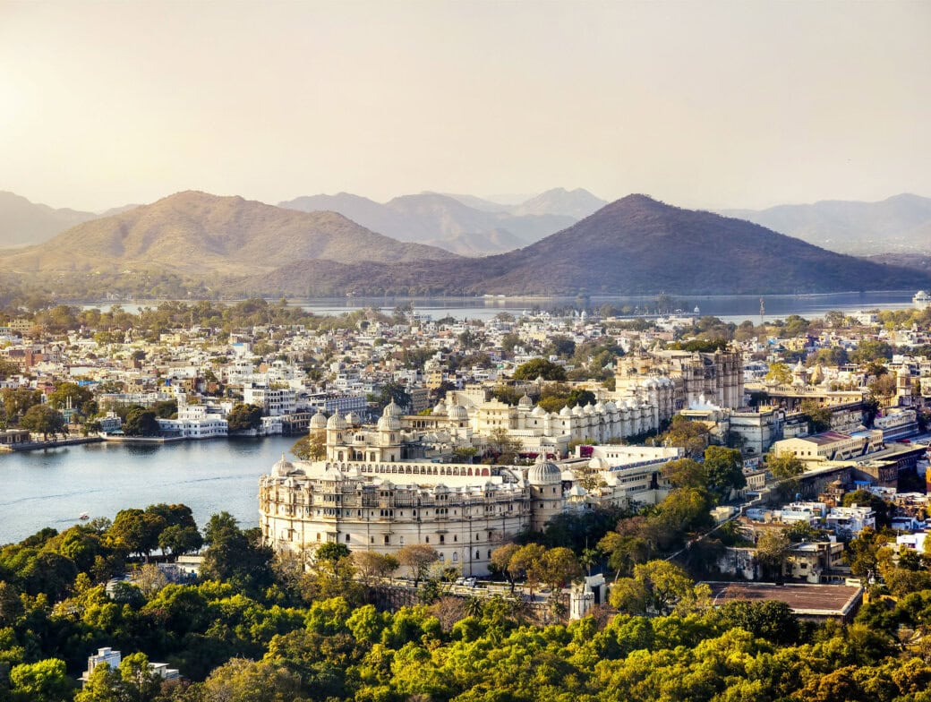 Aerial view of Udaipur, Rajasthan, showcasing historic architecture, lush greenery, a large palace, and distant hills under a hazy sky—a perfect introduction for any beginner's guide to India's beauty.
