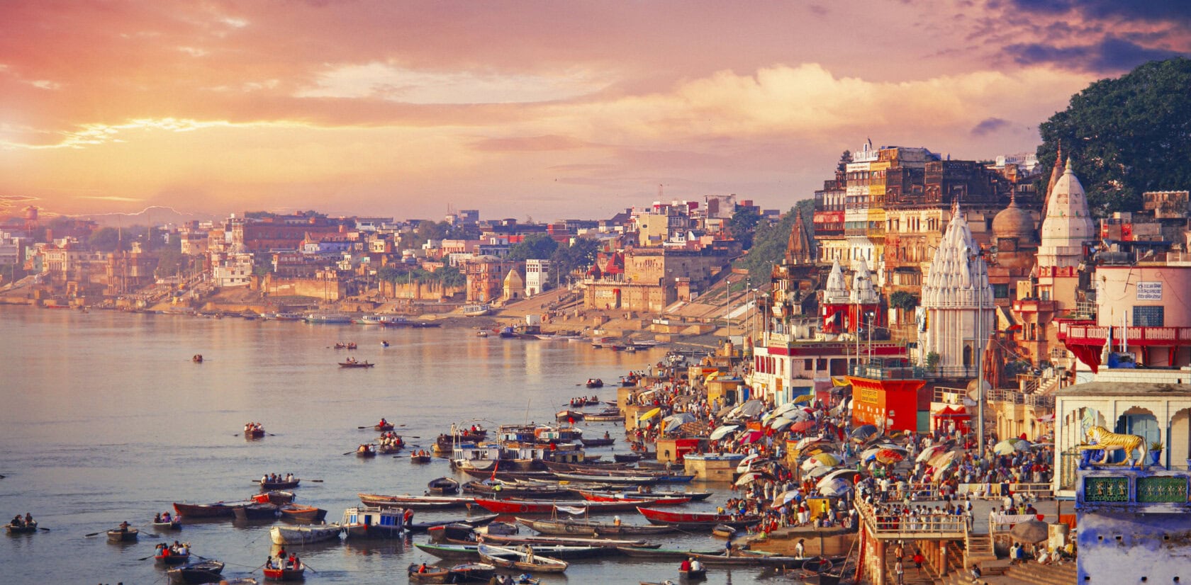 Boats glide along the river in Varanasi, where a crowded shoreline boasts colorful buildings and temples under a dramatic sunset sky. This scene captures the vibrant essence of India.