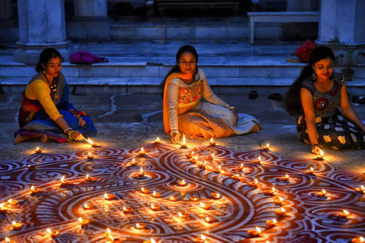 Three women placing oil lamps on a large, intricate rangoli design on the ground, creating an illuminated pattern in a dimly lit environment. Moments like these highlight why India travel is so captivating, offering vibrant cultural experiences throughout your journey.