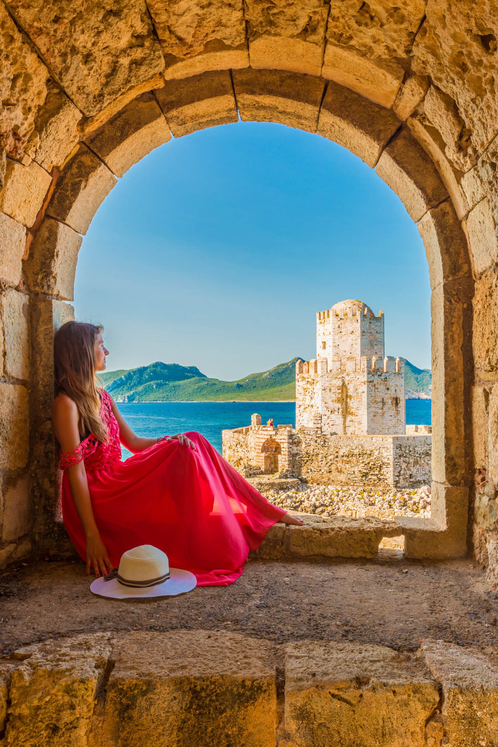 A woman in a red dress and hat from the Cruise Collection sits in an archway, overlooking a historical stone tower. The majestic mountains and blue sky paint the perfect backdrop for this timeless scene.