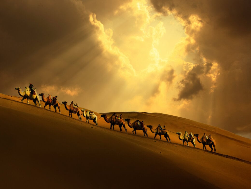 A caravan of camels with riders gracefully traverses a sand dune under a dramatic sky, beams of sunlight breaking through clouds, evoking scenes straight from 2025's Top Ten Destinations list.