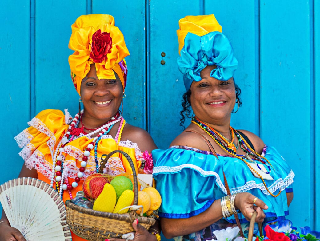 Against a bright blue wall, two women in colorful dresses and headwraps smile warmly, each holding a basket of fruit. One holds a fan as they embrace the vibrant spirit of this Top Ten Destination for 2025 travel enthusiasts.