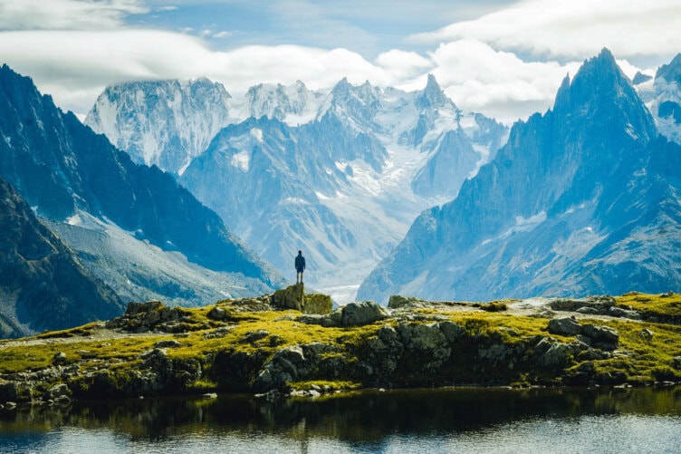 A person stands on a rocky hill overlooking the majestic snow-capped Alps and a clear lake under a cloudy sky, capturing the essence of why this is one of the best countries to visit.