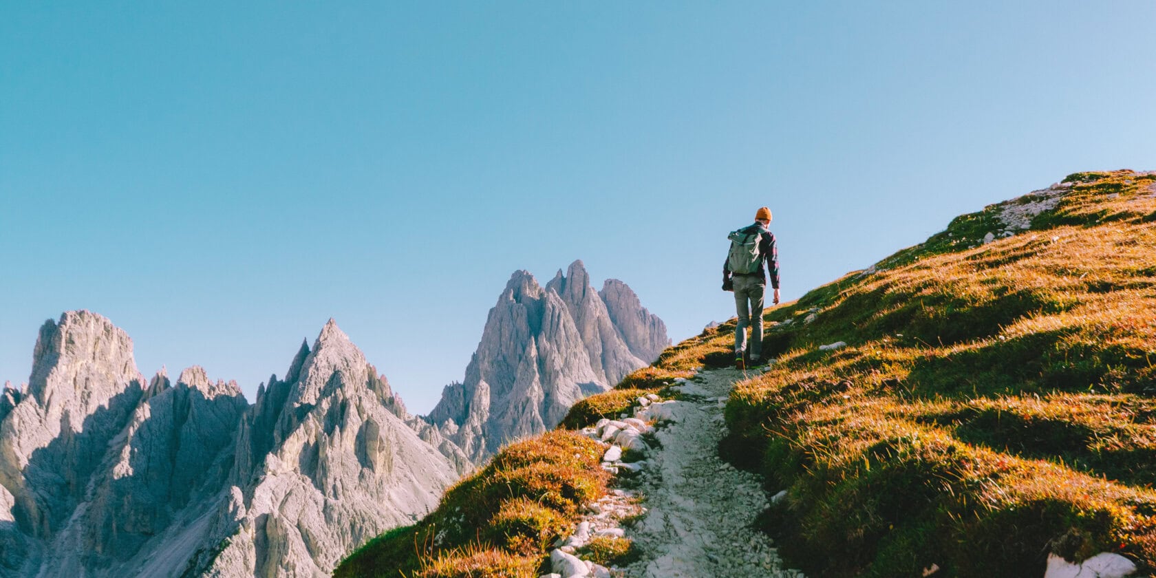 A person hikes a narrow trail on a grassy slope in the Alps, with rocky peaks in the distance under a clear blue sky—a must-visit for those exploring the best countries for breathtaking landscapes.