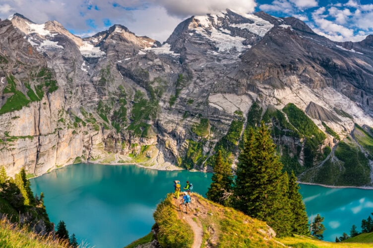 Two hikers stand on a grassy trail in the best country for nature lovers, overlooking a turquoise lake encircled by the towering Alps and dense evergreen trees under a partly cloudy sky.