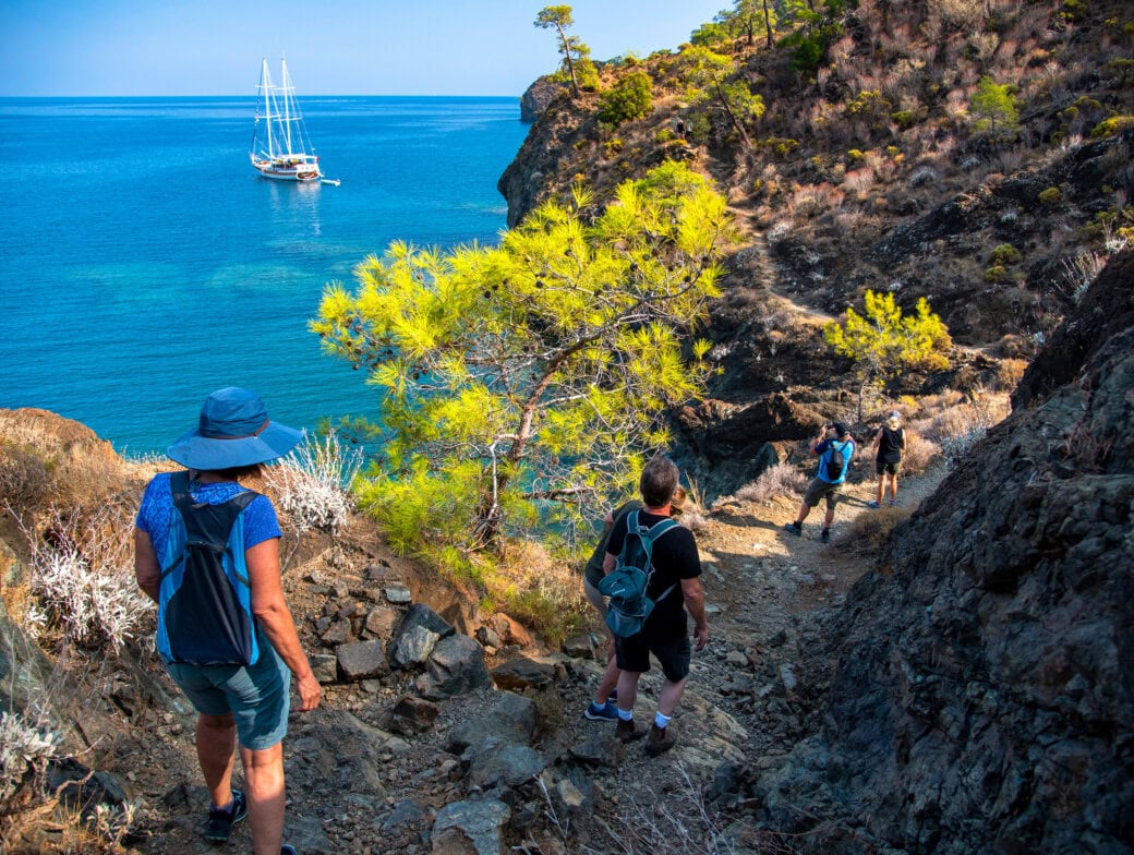 In 2025, four travelers hike down a rocky coastal path towards a sailboat anchored near the shore, with clear blue water and rugged landscape in the background—a scene straight out of the top ten travel destinations.