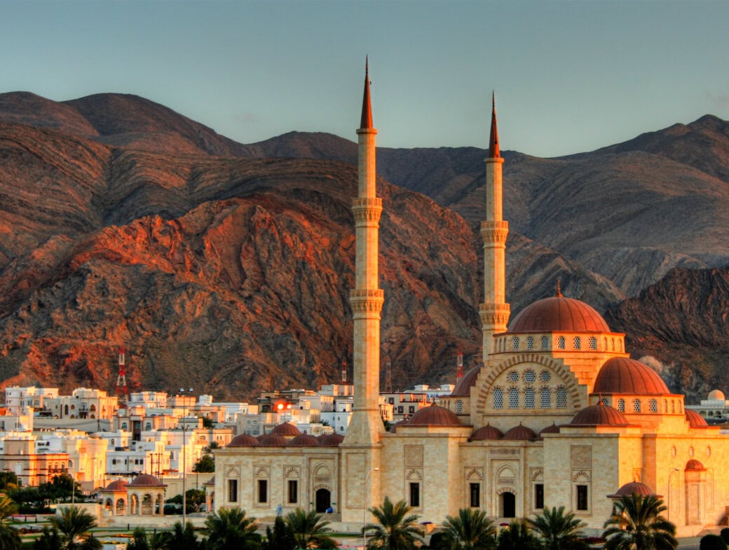 Set against a backdrop of brown mountains and a clear sky, the mosque with its two tall minarets stands prominently, with white buildings in the foreground. As one of the Top Ten Destinations for Travel 2025, it offers an unforgettable experience.