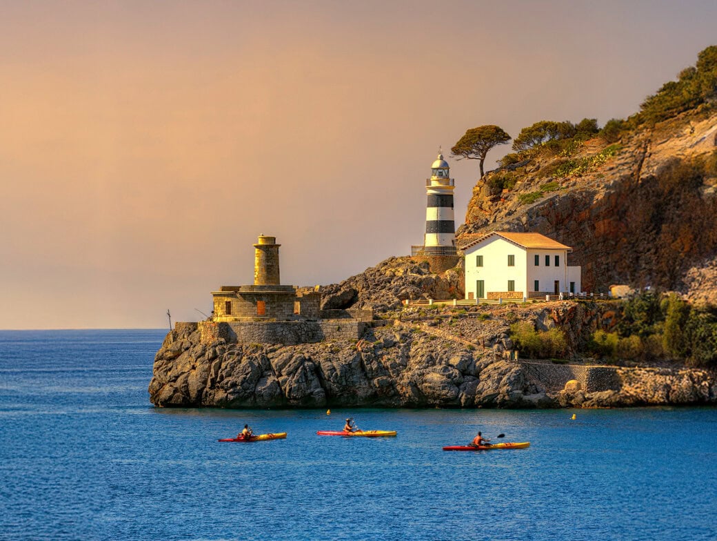 Three kayakers paddle near a rocky coastline featuring a striped lighthouse and a white building under a clear sky, highlighting one of the must-see destinations for travel enthusiasts in 2025.