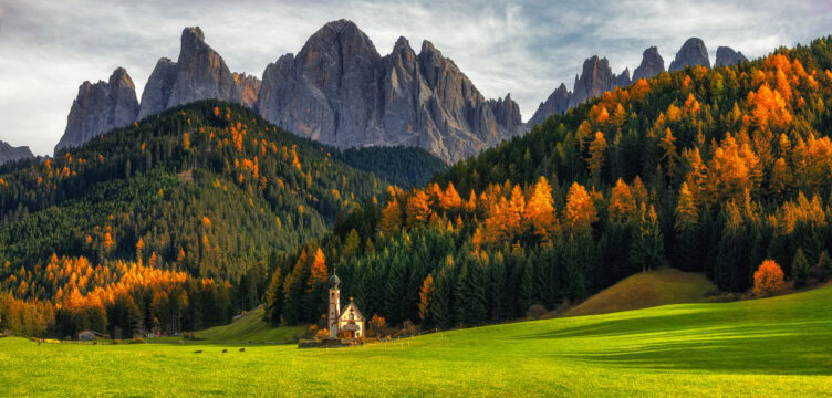 A small church stands in a green field bordered by autumn trees, with the jagged Alps rising majestically in the background under a cloudy sky.