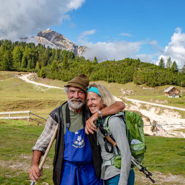An elderly man and woman stand smiling in a grassy landscape with a mountain and trees in the background, embracing their love for travel. The woman, equipped with a backpack and hiking poles, embodies the spirit of adventurous trips taken together.